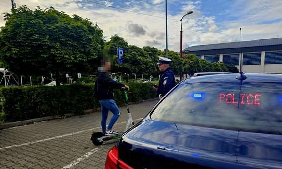 Na pierwszym planie tylna część radiowozu policyjnego, obok policjant i młody chłopak z hulajnogą.