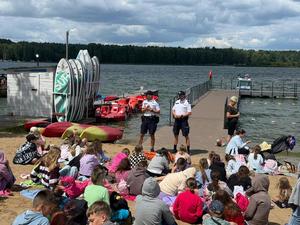 Dwaj policjanci rozmawiają na plaży z grupą dzieci, w tle pomost i akwen wodny.