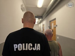 Mężczyzna z policjantem stoją tyłem na korytarzu.