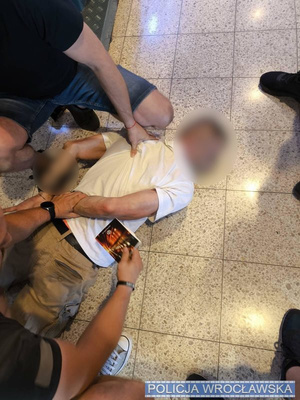 Policjanci przytrzymują leżącego na ziemi zatrzymanego, skutego w kajdanki.
