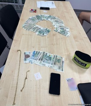 Rozłożone na stole banknoty, dwa telefony komórkowe, opaska z napisem Policja, dwa łańcuszki i paczka papierosów.