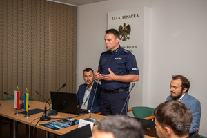 Umundurowany policjant stoi, przy nim siedzą uczestnicy szkolenia.