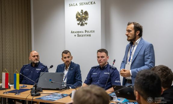 Mężczyzna w garniturze stoi, przy nim siedzą uczestnicy szkolenia.