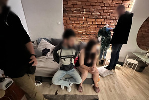 Zatrzymany mężczyzna siedzi na łóżku a przy nim kobieta. Obok stoją dwaj policjanci.