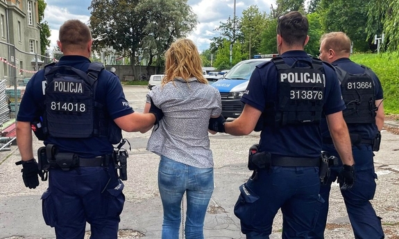 Zatrzymana kobieta prowadzona przez trzech policjantów.
