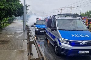 Dwa radiowozu stojące na ulicy, za nimi w kłębach dymu samochód.