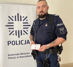 Umundurowany policjant trzyma w rękach legitymację honorowego dawcy krwi. W tle ścianka z napisem: Komenda Miejska Policji w Koszalinie.