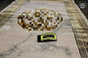 Na podłodze rozwinięty dywan, na którym leży opaska z napisem Policja.