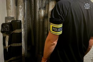 Policjant z opaską na ramieniu Policja, a przed nim stoją przy ścianie zwinięte dywany.