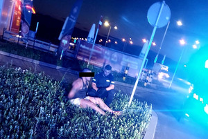 Zatrzymany siedzi zakuty w kajdanki przy drodze na psie zieleni, obok niego kuca policjant.