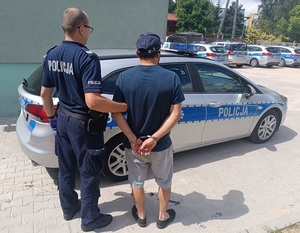 Policjant z zatrzymanym mężczyzną zakutym w kajdanki przy radiowozie.
