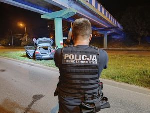 Policjant technik kryminalistyki wykonuje zdjęcia na miejscu wypadku. W głębi samochód.