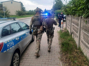 Policjanci na ulicy przed posesją. Wzdłuż ulicy zaparkowane są dwa  radiowozy.