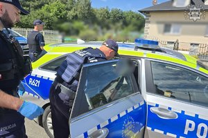 Trzej policjanci przy radiowozie, jeden z nich nachyla się stojąc w drzwiach pojazdu.