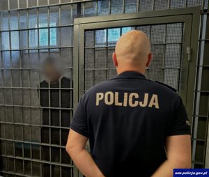 Zatrzymany mężczyzna za kratami i  policjant stojący przed kratami.