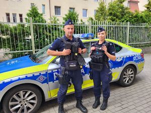 policjant i policjantka stoją obok siebie przed radiowozem.