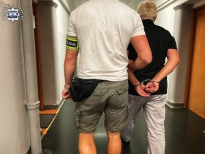 Policjant prowadzi zatrzymanego mężczyznę w kajdankach.