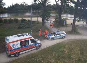 Ratownicy stoją przy radiowozie policyjnym, obok stoi karetka pogotowia. W tle rzeka.