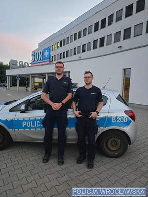 policjanci stoją obok radiowozu.
