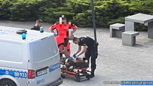 Policjanci i ratownik medyczny z poszkodowanym na noszach przy radiowozie.