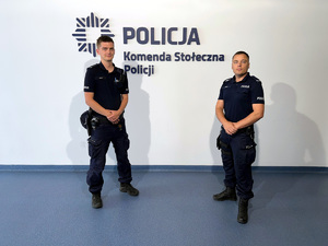 Po lewej st. post. Adrian Tyniec, po prawej sierż. Piotr Jasiński na tle ściany z napisem Komenda Stołeczna Policji.