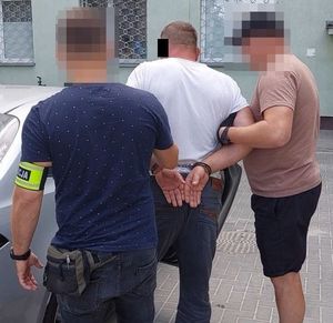 Dwaj funkcjonariusze z zatrzymanym mężczyzną przy nieoznakowanym radiowozie.