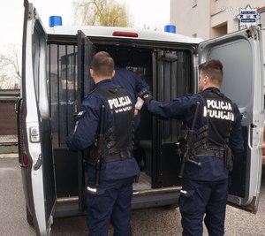 Dwaj policjanci przy radiowozie z otwartymi tylnymi drzwiami. W środku zatrzymany mężczyzna.