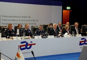 Sala konferencyjna, w której przy długim stole siedzą przedstawiciele w mundurach policyjnych oraz osoby w garniturach. Na stole stoją mikrofony, tabliczki z nazwiskami uczestników oraz karafki z wodą. Zdjęcie przedstawia oficjalne spotkanie w ramach polskiej prezydencji w Radzie Unii Europejskiej.