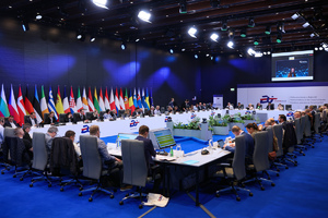 Sala konferencyjna, w której przy stole siedzą przedstawiciele w garniturach. Każda osoba ma przed sobą  laptopa lub dokumenty. Na ścianie w tle widoczny jest rząd flag państwowych ustawionych obok siebie.