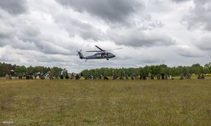 W oddali na polanie grupa policjantów z różnych państw, ustawionych w rzędzie którzy trzymają w ręku flagi różnych państw. Nad nimi śmigłowiec z policjantami.