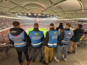 Kryty stadion piłki nożnej z trybunami wypełnionymi kibicami oraz czterech nieumundurowanych policjantów w niebieskich kamizelkach z flagami różnych państw.