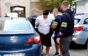 Dwaj policjanci i zatrzymany mężczyzna przy nieoznakowanym radiowozie.
