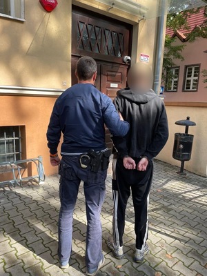Policjant z zatrzymanym mężczyzną zakutym w kajdanki przed budynkiem.