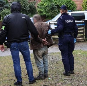 Zdjęcie w kolorze przedstawia parę policjantów prowadzących mężczyznę, który ma założone kajdanki na ręce trzymane z tyłu.W tle brama wjazdowa, za nią radiowóz.