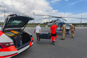 Dwaj mężczyźni niosą skrzynię do śmigłowca. Przy maszynie widać dwóch pilotów.