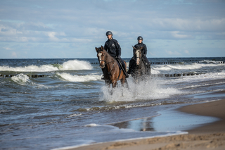Dwoje policjantów na konicha patroluje plażę. Konie rozbryzgują wodę.