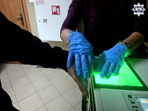Dłoń mężczyzny na urządzeniu do pobierania śladów daktyloskopijnych trzymana przez dłonie w niebieskich rękawiczkach.