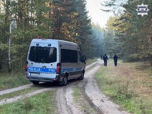 Policjanci idą duktem leśnym, przy którym stoi policyjny radiowóz.