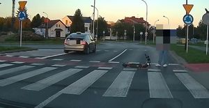 Na zdjęciu przejście dla pieszych. Na lewym pasie widać auto, na prawym pasie mężczyznę z hulajnogą.