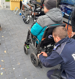 Policjant pomaga przy wózku inwalidzkim, na którym siedzi niepełnosprawny mężczyzna.