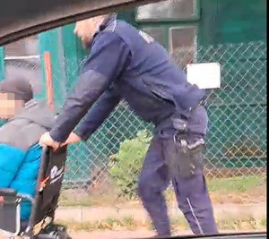 Policjant pcha wózek inwalidzki, na którym siedzi niepełnosprawny mężczyzna.