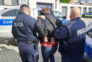 Dwaj funkcjonariusze prowadzą zatrzymanego mężczyznę zakutego w kajdanki.