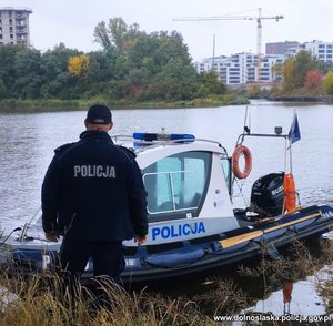 Policjant stoi na brzegu zbiornika wodnego przy zacumowanej łodzi policyjnej.