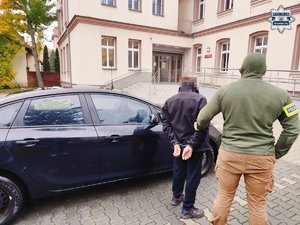 Policjant z zatrzymanym mężczyzną przy radiowozie stojącym przed budynkiem.