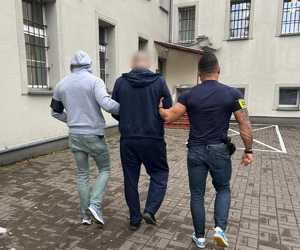 Dwaj policjanci prowadzą zatrzymanego mężczyznę zakutego w kajdanki w stronę budynku.