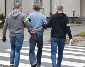 Dwaj funkcjonariusze prowadzą zatrzymanego mężczyznę w kierunku przejścia dla pieszych. W głębi budynek.