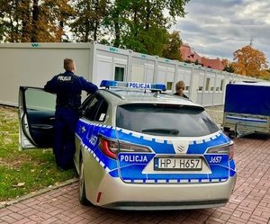 Odwróceni tyłem policjant i policjantka stoją przy radiowozie z otwartymi drzwiami na parkingu przy barakach mieszkalnych.