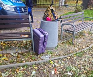 W betonowym koszu na śmieci walizka koloru fioletowego, za koszem policyjny radiowóz i umundurowany policjant.