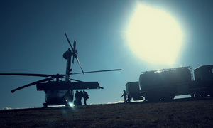 Śmigłowiec stoi na trawiastym lądowisku, obok niego kilka osób oraz samochód ciężarowy z cysterną (zdjęcie wykonane pod słońce).