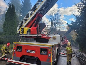 Wóz strażacki i drabina a na wysięgniku strażacy gaszący pożar.